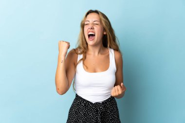 Mavi arka planda izole edilmiş genç sarışın kadın zaferi kutluyor.