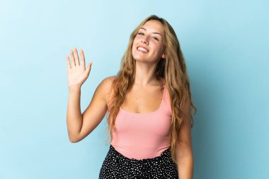 Mavi arka planda izole edilmiş genç sarışın kadın mutlu bir ifadeyle selamlıyor.