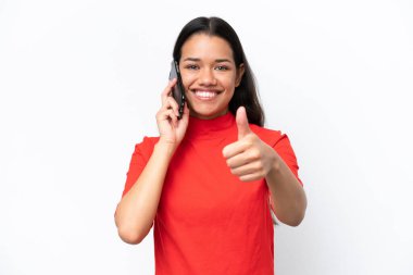 Beyaz arka planda izole edilmiş genç Kolombiyalı kadın başparmaklarını kaldırırken cep telefonuyla sohbet ediyor.