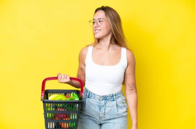 Sarı arka planda izole edilmiş yiyecek dolu bir alışveriş sepeti tutan beyaz bir kadın.