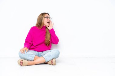 Beyaz arka planda izole bir şekilde yerde oturan genç beyaz kadın ağzı sonuna kadar açık bağırıyor.