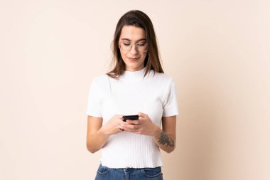 İzole edilmiş bej arka planda genç bir kadın cep telefonuyla mesaj gönderiyor.