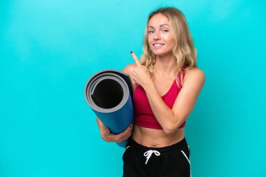 Genç Rus spor kadını yoga derslerine giderken elinde mavi arka planda izole edilmiş bir minder tutarak bir ürünü sunmak için yan tarafa bakıyor.