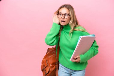 Pembe arka planda izole edilmiş genç bir bayan kulağına elini sürerek bir şeyler dinliyor.