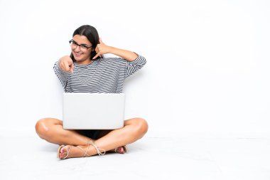 Dizüstü bilgisayarı olan genç, beyaz bir kadın yerde oturuyor telefon hareketleriyle ön tarafı işaret ediyor.