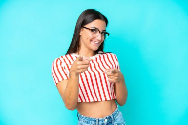 Mavi arka planda izole edilmiş genç beyaz kadın şaşırmış ve ön tarafı işaret ediyor.