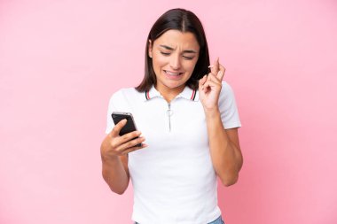 Young caucasian woman isolated on pink background using mobile phone with fingers crossing