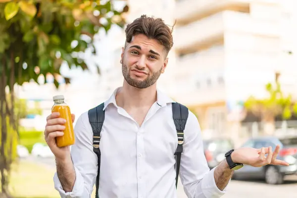 外でオレンジジュースを持っている若いハンサムな男は肩を持ち上げながらジェスチャーを疑う ストック写真