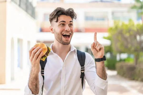 指を上げながら解決策を実現しようとする屋外でハンバーガーを持っている若いハンサムな男 ストック画像