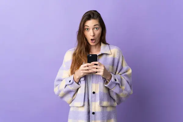 年轻的爱尔兰女人被紫色背景隔离 一边看着相机 一边用手机 脸上带着惊讶的表情 免版税图库图片