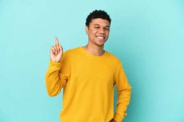 Genç Afro-Amerikan adam mavi arka planda izole edilmiş ve en iyinin işareti olarak parmağını kaldırıyor.