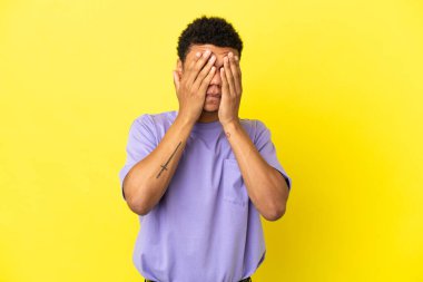 Genç Afrikalı Amerikalı adam sarı arka planda izole edilmiş gözleri elleriyle kapatıyor.