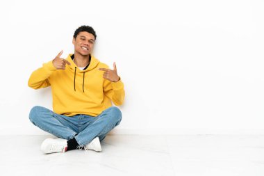 Genç Afrikalı Amerikalı bir adam beyaz arka planda soyutlanmış gururlu ve kendini beğenmiş bir şekilde yerde oturuyor.