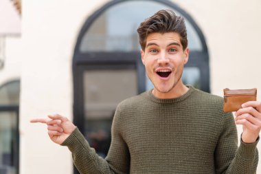 Açık havada cüzdan tutan genç, beyaz bir adam şaşırmış ve parmağıyla onu işaret ediyor.