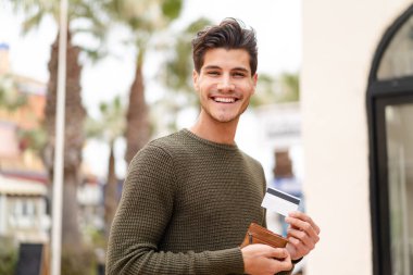 Açık havada genç beyaz bir adam elinde cüzdan ve mutlu bir ifadeyle kredi kartı tutuyor.