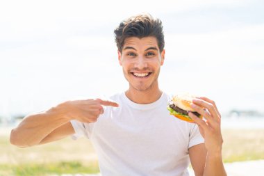 Genç, beyaz bir adam elinde sürpriz bir yüz ifadesiyle açık havada hamburger tutuyor.