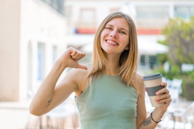 Genç sarışın kadın açık havada kahvesini yudumlarken gururlu ve kendinden memnun.