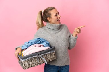 Genç bir kadın pembe arka planda izole edilmiş bir çamaşır sepetini tutuyor. Parmakla göstererek bir ürün sunuyor.