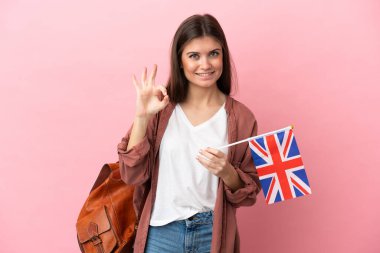 Pembe arka planda izole edilmiş bir Birleşik Krallık bayrağı tutan genç beyaz kadın parmaklarıyla tamam işareti gösteriyor.