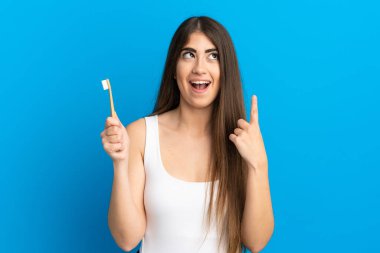 Dişlerini fırçalayan beyaz bir kadın mavi arka planda izole edilmiş bir şekilde parmak kaldırmayı düşünüyor.