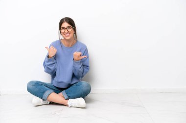 Beyaz arka planda izole bir şekilde yerde oturan beyaz bir kadın. Baş parmakları havada ve gülümsüyor.