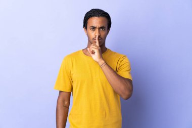 Örgülü genç Afrikalı Amerikalı adam mor arka planda izole edilmiş. Parmağını ağzına koyan sessizlik işareti gösteriyor.