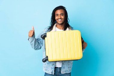 Saçları mavi arka planda izole edilmiş, seyahat valiziyle gezen genç Afrikalı Amerikalı bir adam.