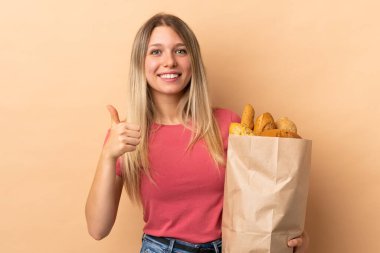 Elinde bej arka planda izole edilmiş bir çanta dolusu ekmek tutan genç sarışın kadın baş parmak işareti yapıyor.