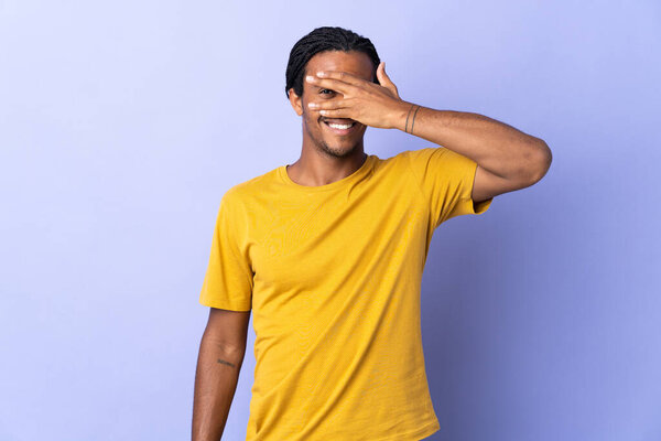 Young African American man with braids man isolated on purple background covering eyes by hands and smiling