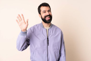 Sakallı, ceketli, soyutlanmış arka planda beyaz bir adam mutlu bir ifadeyle selam veriyor.