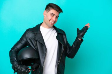 Motosiklet kasklı genç beyaz adam mavi arka planda izole edilmiş. Davet etmek için ellerini yana uzatıyor.