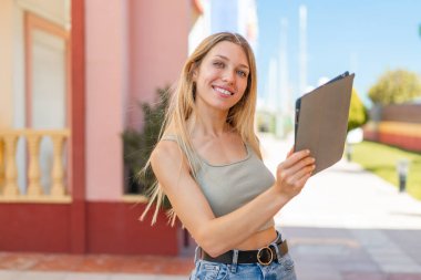 Açık havada elinde mutlu bir ifadeyle tablet tutan genç sarışın kadın.