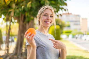 Sarışın bir kadın portakalı açık havada tutuyor ve ona doğrultuyor.