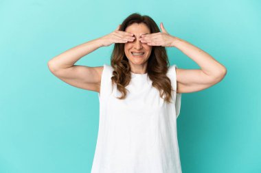 Orta yaşlı beyaz kadın mavi arka planda izole edilmiş gözleri elleriyle kaplayan ve gülümseyen.