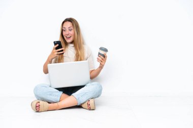 Young woman with laptop sitting on the floor isolated on white background holding coffee to take away and a mobile