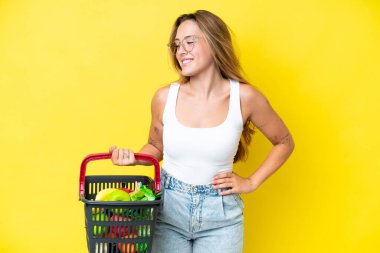 Elinde yiyecek dolu bir sepet taşıyan beyaz bir kadın. Sarı arka planda izole edilmiş. Kolları kalçasında ve gülümsüyor.