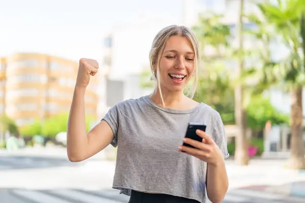 Jeune Femme Blonde Extérieur Utilisant Téléphone Mobile Faisant Geste Victoire Images De Stock Libres De Droits