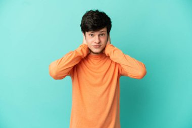 Mavi arka planda izole edilmiş genç Rus adam hayal kırıklığına uğramış ve kulaklarını kapatmış.