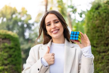 Genç ve güzel bir kadın dışarıda üç boyutlu bir bulmaca küpünü tutuyor. Başparmakları havada, çünkü iyi bir şey oldu.
