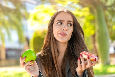 Açık havada elinde elmayla donut tutan genç bir kadın ve şüpheleri var.