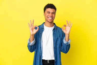 Sarı arka planda izole edilmiş genç Afrikalı Amerikalı iki eliyle tamam işareti gösteriyor.