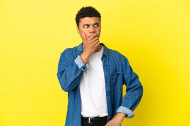 Genç Afro-Amerikalı adam sarı arka planda izole edilmiş. Şüpheleri var ve kafası karışmış bir yüz ifadesi var.