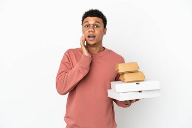 Elinde hamburger ve pizza tutan genç Afrikalı bir adam beyaz arka planda şaşırmış ve şok olmuş bir yüz ifadesi ile izole edilmiş.