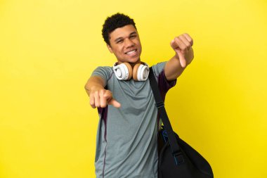 Sarı arka planda izole edilmiş spor çantalı genç Afrikalı Amerikalı adam gülümserken parmağını sana gösteriyor.