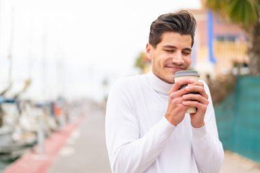 Genç beyaz bir adam açık havada elinde kahve tutuyor.