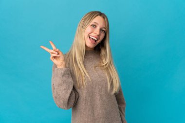 Mavi arka planda izole edilmiş genç sarışın kadın gülümsüyor ve zafer işareti gösteriyor.