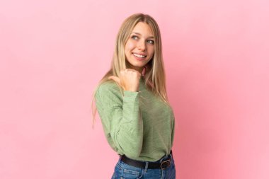 Pembe arka planda izole edilmiş genç sarışın kadın bir ürünü sunmak için kenarı işaret ediyor.