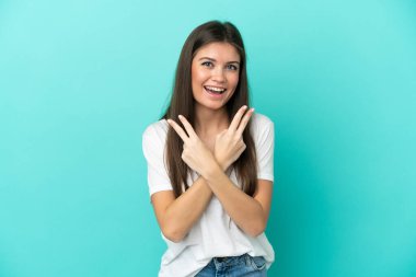 Mavi arka planda izole edilmiş genç beyaz kadın gülümsüyor ve zafer işareti gösteriyor.