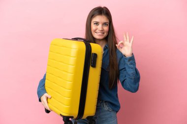 Genç beyaz kadın pembe arka planda izole edilmiş seyahat valiziyle geziyor ve 