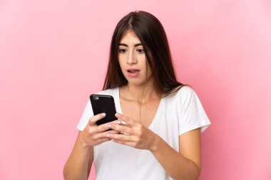 Pembe arka planda izole edilmiş genç beyaz kadın kameraya bakarken şaşırmış bir ifadeyle cep telefonunu kullanıyor.